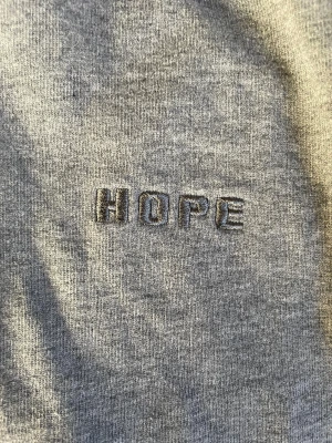 Grå hoodie från HOPE Stockholm - Snygg grå hoodie från HOPE Stockholm med broderad logga på bröstet. Klassisk känguruficka framtill och huva. Perfekt för en chill och stilren look. Tillverkad i mjuk bomull för extra komfort.