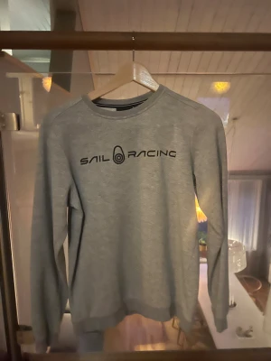 Grå Sail Racing sweatshirt - Snygg grå sweatshirt från Sail Racing med svart logga tryckt på bröstet. Tröjan har rund halsringning, långa ärmar och ribbade muddar vid ärmslut och nederkant. Perfekt för dig som gillar en clean och sportig stil.