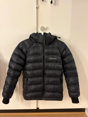 Svart Peak Performance dunjacka - Svart dunjacka från Peak Performance med huva och broderad logga på bröstet. Jackan har quiltad design, dragkedja framtill och muddar vid ärmslut. Perfekt för kalla dagar och har en clean, sportig look som funkar till det mesta.