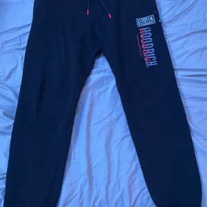 Svarta Hoodrich mjukisbyxor med tryck - Svarta mjukisbyxor från Hoodrich med vitt och rött tryck längs benet och coola streetwear-detaljer. Byxorna har snörning i midjan och ribbade muddar vid benslut. Perfekta för chill eller streetstyle, med skön passform och mjukt material.