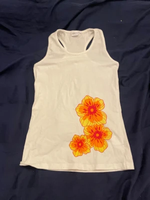 Vit linnetopp med blommor från Mywear - Vit ärmlös linnetopp från Mywear med stora, färgglada blommor i orange och rött tryckta framtill. Modellen har brottarrygg och är gjord i mjuk bomull, perfekt för varma dagar och en somrig look.