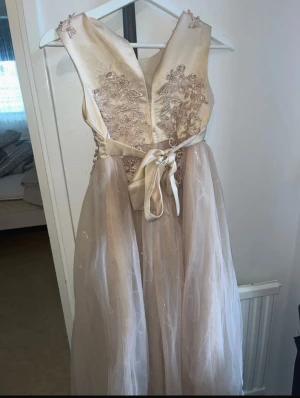 Barn långklänning  - Elegant långklänning i champagnefärg med vackra broderade blommor och pärldetaljer på livet. Klänningen har ett satinliknande överdel, markerad midja med knytband och ett voluminöst tyllkjol. Perfekt för bal eller speciella tillfällen.