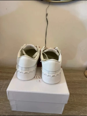 Valentino Rockstud vita sneakers - Säljer ett par vita Valentino Rockstud sneakers i skinn med ikoniska nitar i ljusrosa längs sidorna och bak på skon. Klassisk rund tå, platt sula och snörning framtill. Snygg och stilren design som sticker ut med de unika detaljerna.