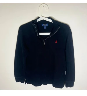 Svart stickad half zip från Polo Ralph Lauren - Svart stickad tröja från Polo Ralph Lauren med röd broderad logga på bröstet. Tröjan har half zip-dragkedja i halsen och långa ärmar med ribbade muddar. Perfekt för dig som gillar klassisk och stilren design.