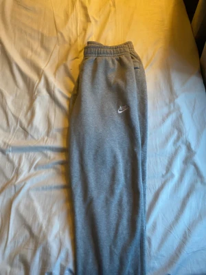 Grå mjukisbyxor från Nike, storlek L - Grå mjukisbyxor från Nike med broderad logga på vänster ben. Byxorna har elastisk midja och muddar vid bensluten för skön passform. Tillverkade i mjukt bomullsmaterial, perfekta för chill eller träning. Klassisk och enkel design som alltid funkar.
