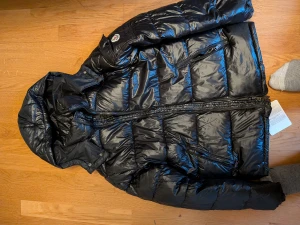 Svart Moncler pufferjacka med huva - Svart pufferjacka från Moncler med huva och quiltad design. Jackan har dragkedja framtill, praktiska fickor med dragkedja och en ikonisk Moncler-logga på axeln. Insidan har en cool serietidningsetikett och märkning från DIST. Perfekt för dig som vill ha en varm och snygg jacka.