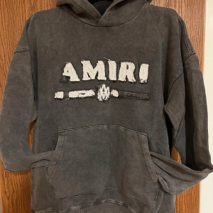 Grå hoodie från Amiri med broderi - Säljer en grå hoodie från Amiri med stor broderad logga i vitt och svart på bröstet. Tröjan har huva, magficka och långa ärmar. Materialet är mjukt och känns som bomull, med en tvättad vintage-look. Perfekt för dig som gillar streetwear och snygga detaljer.