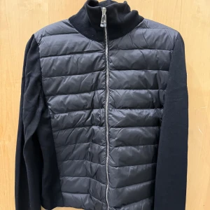 Svart pufferjacka från Moncler - Svart pufferjacka från Moncler med quiltad front och rygg i glansig polyester, samt stickade ärmar och krage. Jackan har dragkedja framtill och Moncler-logga på ena ärmen. Perfekt för dig som vill ha en stilren och sportig look.