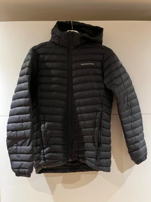 Svart dunjacka Peak Performance - Säljer en svart dunjacka från Peak Performance med huva och dragkedja framtill. Jackan har quiltad design och två fickor med dragkedja. Perfekt för kalla dagar och riktigt skön att bära. Klassisk och stilren look.