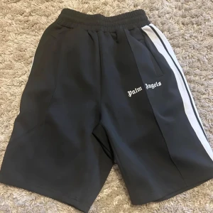 Palm Angels svarta shorts med vit stripe - Säljer ett par svarta shorts från Palm Angels med vit stripe längs sidorna och logga tryckt på vänster ben. De har elastisk midja och en loose fit som ger en avslappnad vibe. Perfekta för sommaren och streetwear-stilen.