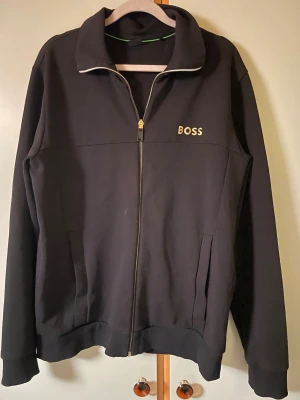 Svart zipjacka från BOSS, storlek M - Svart zipjacka från BOSS med guldbroderad logga på bröstet. Jackan har ståkrage, hel dragkedja och två sidofickor. Materialet är mjukt och känns som bomull eller polyester, perfekt för chill dagar. Snygg och enkel design med ribbade muddar. Aldrig använt vill få bort den för den tar plats 