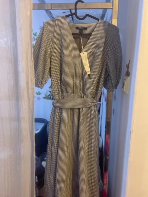 Rutig långklänning från Esprit - Supersnygg långklänning från Esprit i svartvitt rutigt mönster. Klänningen har v-ringad omlottfront, puffiga kortärmar och ett markerat midjeband. Perfekt för dig som gillar klassisk stil med en modern twist. Materialet är mjukt och känns skönt mot huden.