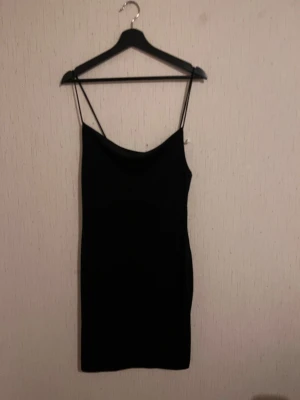 Svart kort klänning med smala axelband - En stilren svart kort klänning med smala axelband och rak passform. Klänningen har en enkel och klassisk design med lätt draperad urringning. Perfekt för dig som gillar minimalistisk stil och vill ha något som känns både trendigt och tidlöst.