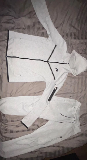 Grå Nike tech fleece Hoodie och Byxor - Snygg grå hoodie från Nike med svarta dragkedjor och detaljer. Tröjan har huva, lång ärm och en cool svart linje över bröstet. Perfekt för chill eller träning, med Nike-logga på bröstet och ärmen. Mjuk bomullskänsla och normal passform.