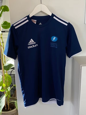Marinblå Adidas fotbollströja SFA! NY!  - Säljer en marinblå fotbollströja från Adidas med Svenska Fotbollsakademin-tryck och Stadium-logga. Tröjan har vita detaljer, klassiska Adidas-ränder på axlarna och är gjord i lätt, ventilerande polyester. Kortärmad modell med rund hals och Climalite-material.