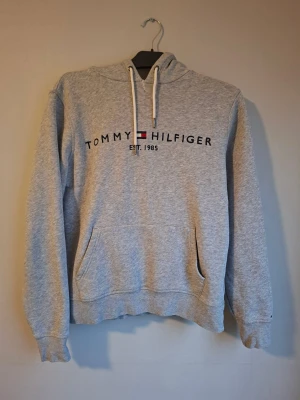 Grå hoodie Tommy Hilfiger - Snygg grå hoodie från Tommy Hilfiger med broderad logga och text 'EST. 1985' på bröstet. Tröjan har huva med vita snören, magficka och långa ärmar. Perfekt för chill dagar och enkel att matcha med jeans eller joggers. Har sytt lite uppe som man ser på bilden men de syns knappt!