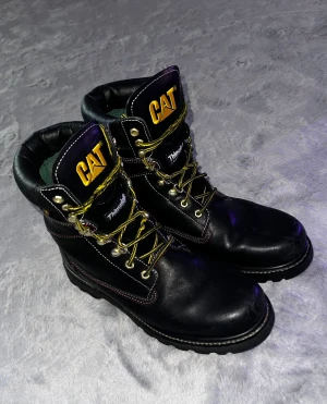 Svarta CAT boots med gul logga - CAT boots i svart skinn med grov rödsvart sula och gul logga på plösen. Skorna har gula snören, metallöglor och röd kontrastsöm. Insidan är fodrad med Thinsulate för extra värme. Perfekt för dig som gillar robust stil och vill sticka ut.