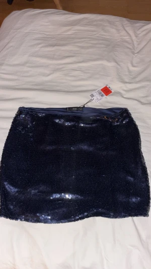 Mörkblå paljettkjol från MNG - Supersnygg mörkblå kortkjol från MNG, täckt med glittrande paljetter som ger en riktigt festlig vibe. Kjolen har en rak passform och är fodrad för extra komfort. Perfekt för dig som vill sticka ut med en glammig look och nyår