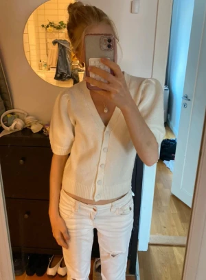 Vit stickad kofta från Gina Tricot - Supersöt vit stickad kofta från Gina Tricot med puffiga korta ärmar och v-ringning. Koftan har ribbade muddar och knappar framtill, perfekt för lager-på-lager och chill stil. Passar dig som gillar en clean och fräsch look. Tröjan är helt ny endast testad så den är i superfint skick!