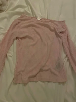 Ljusrosa ribbad offshoulder topp - Säljer en ljusrosa ribbad topp med offshoulder-design och långa ärmar. Toppen har en mjuk och stretchig känsla, perfekt för att skapa en chill och snygg look. Passformen är normal och materialet känns bekvämt mot huden.