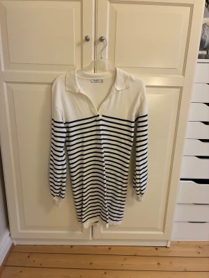 Randig finstickad klänning från NA-KD - Snygg finstickad klänning från NA-KD i vit med marinblå ränder. Tröjan har krage, v-ringning och långa ärmar med ribbade muddar. Materialet är mjukt och skönt stickat. Storlek M. Oanvänd, endast tvättad.