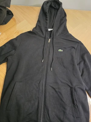 Lacoste - Svart hoodie från Lacoste i storlek M med klassisk krokodillogga på bröstet. Tröjan har huva med snörning, två fickor fram och hel dragkedja. Tillverkad i mjuk bomull, perfekt för chill dagar. Snygg och enkel design som funkar året runt.