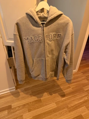 Grå hoodie med dragkedja Maggiore - Grå hoodie från Maggiore med broderad logga framtill. Tröjan har huva, två stora fickor och dragkedja hela vägen. Perfekt för chill dagar och enkel att matcha med jeans eller joggers. Mjuk och skön polyesterblandning.