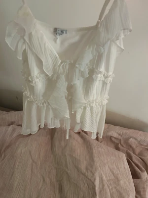 Vit volangig blus med knyt  - Supersöt vit blus med tunna axelband, volanger och plisserade detaljer. Blusen har knyt framtill och flera lager volanger som ger en romantisk vibe. Perfekt för dig som gillar en luftig och feminin stil.