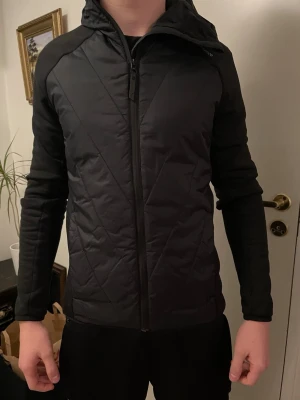 Svart vindjacka Peak Performance S - Svart hybridjacka från Peak Performance i storlek S. Personen på bilden är 179 cm lång. Jackans kantband är sytt men inget som syns tydligt vid användning och knappt när man kollar noga. Jackan har ett nypris på ca 2500 kr. Hör av er vid frågor!
