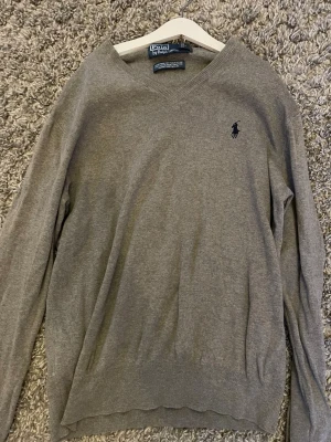 Grå v-ringad tröja från Polo Ralph Lauren - Snygg grå v-ringad tröja från Polo Ralph Lauren i mjuk pima bomull. Klassisk design med mörkblå broderad logga på bröstet och ribbade muddar vid ärmslut och nederkant. Perfekt för dig som gillar stilrena och tidlösa plagg.