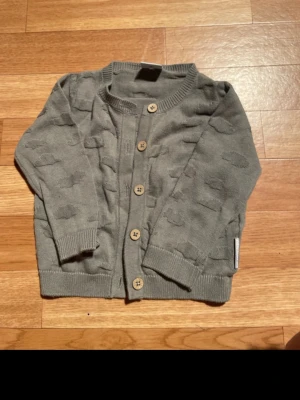 Grå kofta från Polarn O. Pyret - Säljer en grå kofta från Polarn O. Pyret i storlek 68 för bebisar 4-6 månader. Koftan har ett diskret mönster, ribbade muddar och träknappar framtill. Materialet är mjukt och stickat, perfekt för kyliga dagar.