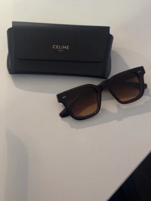 CHIMI CORE 04 solglasögon - Säljer ett par snygga solglasögon från CHIMI, modell CORE 04 i brun färg. Glasögonen har en fyrkantig form och bruna tonade linser. Designade i Stockholm och kommer med ett svart fodral från Celine. Perfekt accessoar för soliga dagar. Nytt skick!