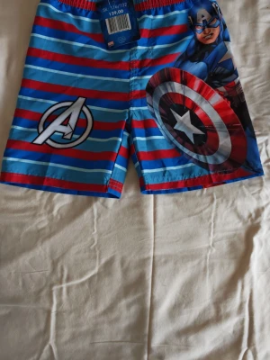 Barn badshorts str.122/128 Marvel Avengers - Snygga badshorts från Marvel med Avengers-tryck och Captain Americas sköld. I str 122/128.Shortsen är randiga i blått, rött och ljusblått och har resår i midjan. Tillverkade i 100% polyester med innerbyxa i mesh.  för stranden eller poolen!