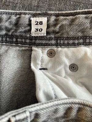 Grå jeans från Jack & Jones, storlek 28/30 - Säljer ett par grå jeans från Jack & Jones i klassisk femficksmodell. Jeansen har en rak passform och är tillverkade i slitstarkt denimtyg med ljusgrå tvätt. Fint skick förutom en fläck på framsidan av byxa(se bild). Strl 28/30