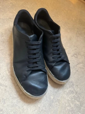 Axel arigatos storlek 45 - Svarta sneakers i skinn med rund tå och platt vit sula. Skorna har klassisk snörning och en stilren design utan synliga loggor. Perfekt för dig som gillar minimalistisk stil och vill ha ett par enkla, svarta sneakers som funkar till det mesta.