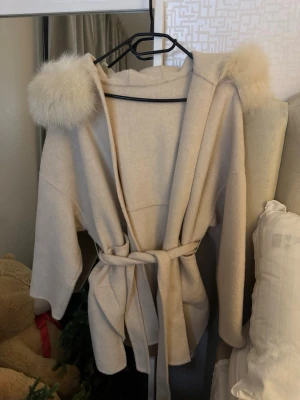 Creamvit kappa med pälsdetaljer och bälte - Säljer en Creamvit kappa med fluffiga pälsdetaljer på huvan och ett snyggt knytbälte i midjan. Jätte fint skick inga defekter🤍