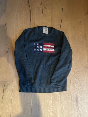 Mörkblå stickad Lexington tröja S - Säljer en mörkblå stickad tröja från Lexington med broderad flaggdetalj och texten 'New Eng. Est. 1993' på bröstet. Tröjan har rund halsringning, ribbade muddar och lång ärm. Perfekt för dig som gillar klassisk college-stil.