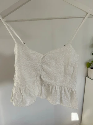 Vit spetsig topp med volang - Vit topp med smala justerbara axelband och volang nedtill. Toppen har broderade spetsdetaljer och ett smockat parti baktill för extra komfort. Från märket HouseBrand i stl s, kan även passa xs.