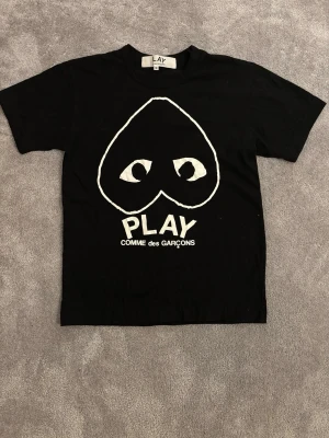 Svart t-shirt Comme des Garçons Play - Svart t-shirt från Comme des Garçons Play med stort vitt tryck på bröstet som föreställer ett hjärta med ögon och texten PLAY. Klassisk rund hals och korta ärmar. Materialet är mjuk bomull som känns skönt mot huden.