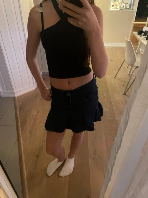 Mörkblå jeanskjol med volang - Säljer en mörkblå jeanskjol med volangkant och råa kanter nertill. Kjolen har klassisk femficksdesign och stängs med knapp och dragkedja framtill. Perfekt för dig som gillar en trendig och avslappnad stil.