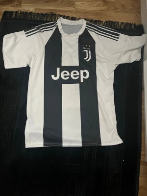 Juventus fotbollströja Ronaldo #7 - Juventus matchtröja med klassiska svarta och vita ränder, Adidas-logga och Jeep som sponsor. På ryggen står Ronaldo och nummer 7. Tröjan har korta ärmar och är gjord i polyester, perfekt för fotbollsfans.