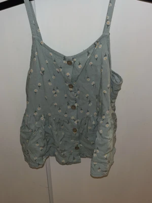 Blommig linnetopp från H&M - Söt ljusgrön linnetopp från H&M med små vita och gula blommor. Toppen har knappar framtill, volang nedtill och smala axelband. Perfekt för varma dagar och enkel att matcha med jeans eller shorts.