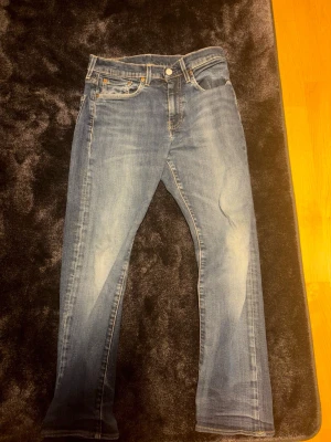 Levi's 502 blå jeans, W28 L32 - Säljer ett par Levi's 502 jeans i klassisk blå tvätt med snygga slitningar. Modellen har rak passform, fem fickor och bruna detaljer på läderpatchen bak. Jeansen är gjorda i mjukt bomullsmaterial och har normal midja. Perfekt för dig som gillar tidlös denimstil.