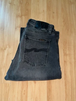Nudie Jeans - Säljer ett par mörk gråa Nudie Jeans. Jeansen har hög midja och raka ben. Midja 30cm, längd 102cm. JAG SKICKAR EJ FLER BILDER NÄR JAG BÄR JEANSEN! för bättre inblick i passformen rekommenderar jag att googla på modellnamnet. För storleksguide kolla måtten noggrant då jeansen alltid kan vara uppsydda heller krympta och jag ej tar emot returer :) s5,4