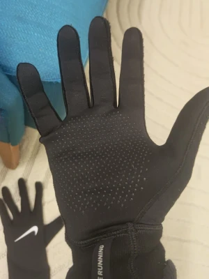 Svarta Nike löparhandskar - Säljer ett par svarta Nike löparhandskar med vit logga på ovansidan. Handskarna har greppvänliga prickar i handflatan och är tillverkade i ett stretchigt syntetmaterial som passar perfekt för träning utomhus. Fungerar att använde på telefon. Storlek L, till en viss del vattentäta. Fungerar jätte bra för varje dag använding på vintern