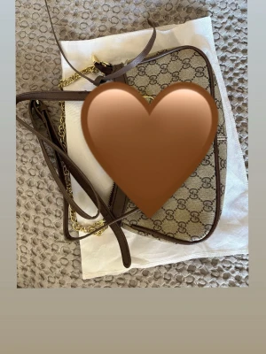  brun axelväska med kedja - Säljer en snygg axelväska  med klassiskt GG-mönster i beige och brunt. Väskan har detaljer i mörkbrunt skinn, guldfärgad kedja och dragkedja upptill. Perfekt storlek för det viktigaste och har en stilren form med justerbar axelrem.