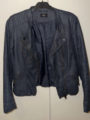 Svart skinnjacka från ONLY - Snygg svart skinnjacka från ONLY med coola detaljer som snedställda dragkedjor och markerade axlar. Jackan har lång ärm och klassisk bikerstil som ger en edgy vibe. Perfekt för dig som vill ha en stilren och trendig look.