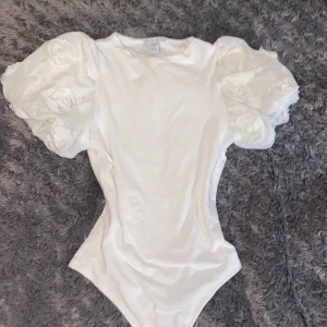 Vit blus med puffärmar från Amisu - Säljer en vit blus från Amisu i storlek S. Toppen har stora puffärmar dekorerade med tygrosor som ger en extra cool och romantisk vibe. Kroppen är figurnära och slät, perfekt för att sticka ut. Materialet känns mjukt och har en lätt glans.