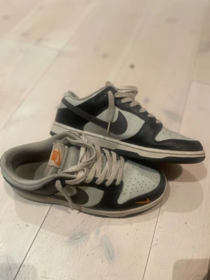 Nike Dunk Low svart och grå EUR 42 - Nike Dunk Low sneakers i svart och ljusgrått med orange Nike-logga på tungan och vid tåspetsen. Skorna har snörning, rund tå och platt sula. Tillverkade i skinn och syntetmaterial med perforerade detaljer på ovansidan för extra ventilation.