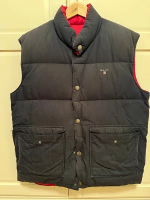 Mörkblå/röd vändbar dunväst från GANT - Säljer en mörkblå/röd vändbar dunväst från GANT. Västen har två stora fickor framtill och hög krage. GANT-logga broderad på bröstet.